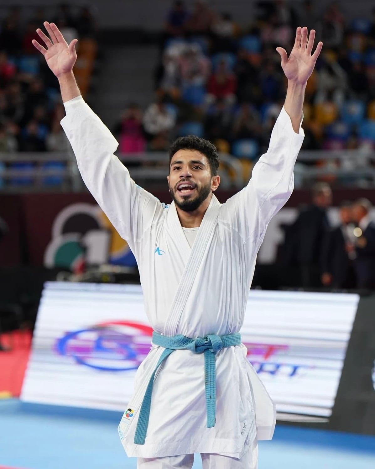 Abdallah Hesham with both arms raised after a win, the competition arena filled behind him.