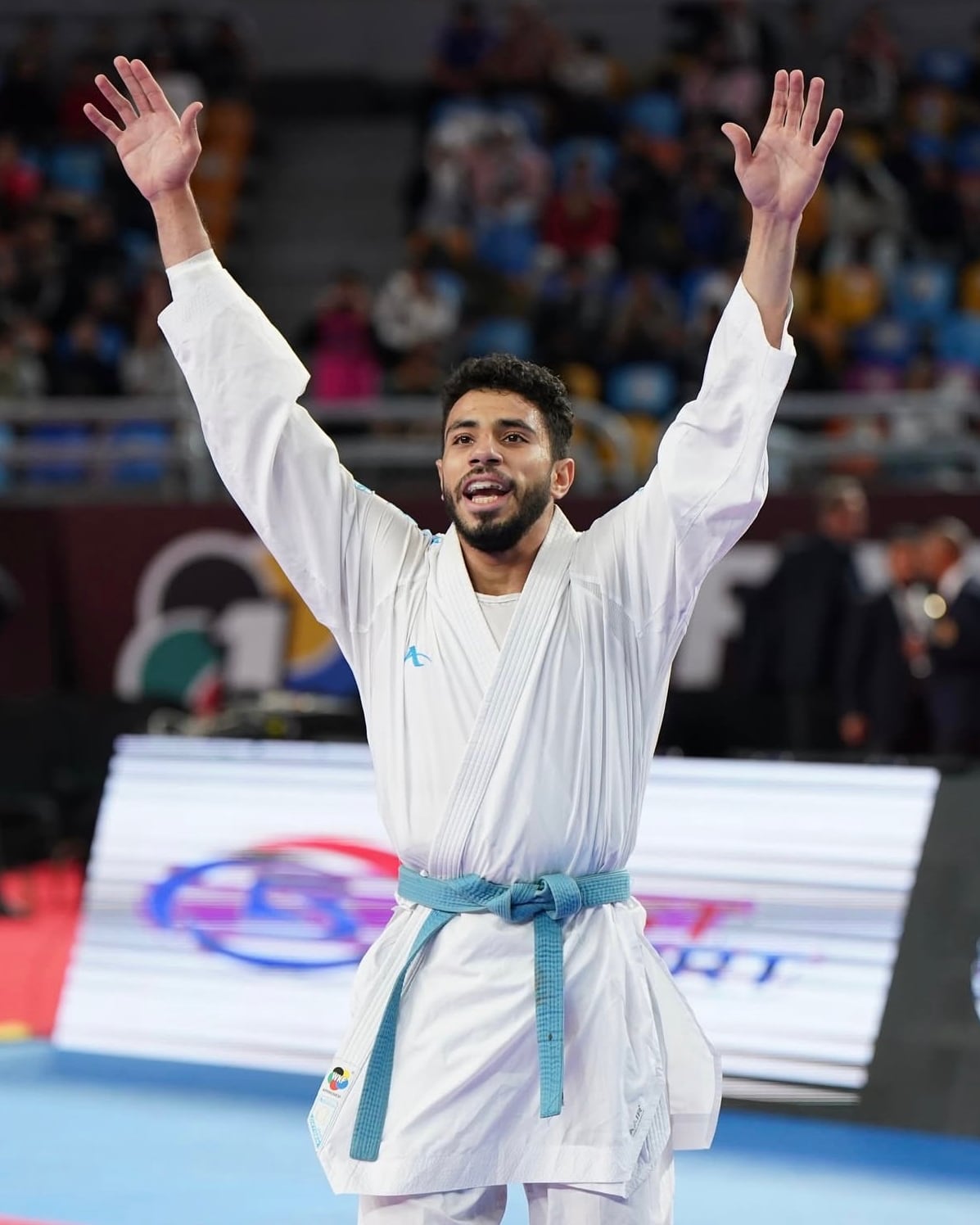 Abdallah Hesham with both arms raised after a win, the competition arena filled behind him.