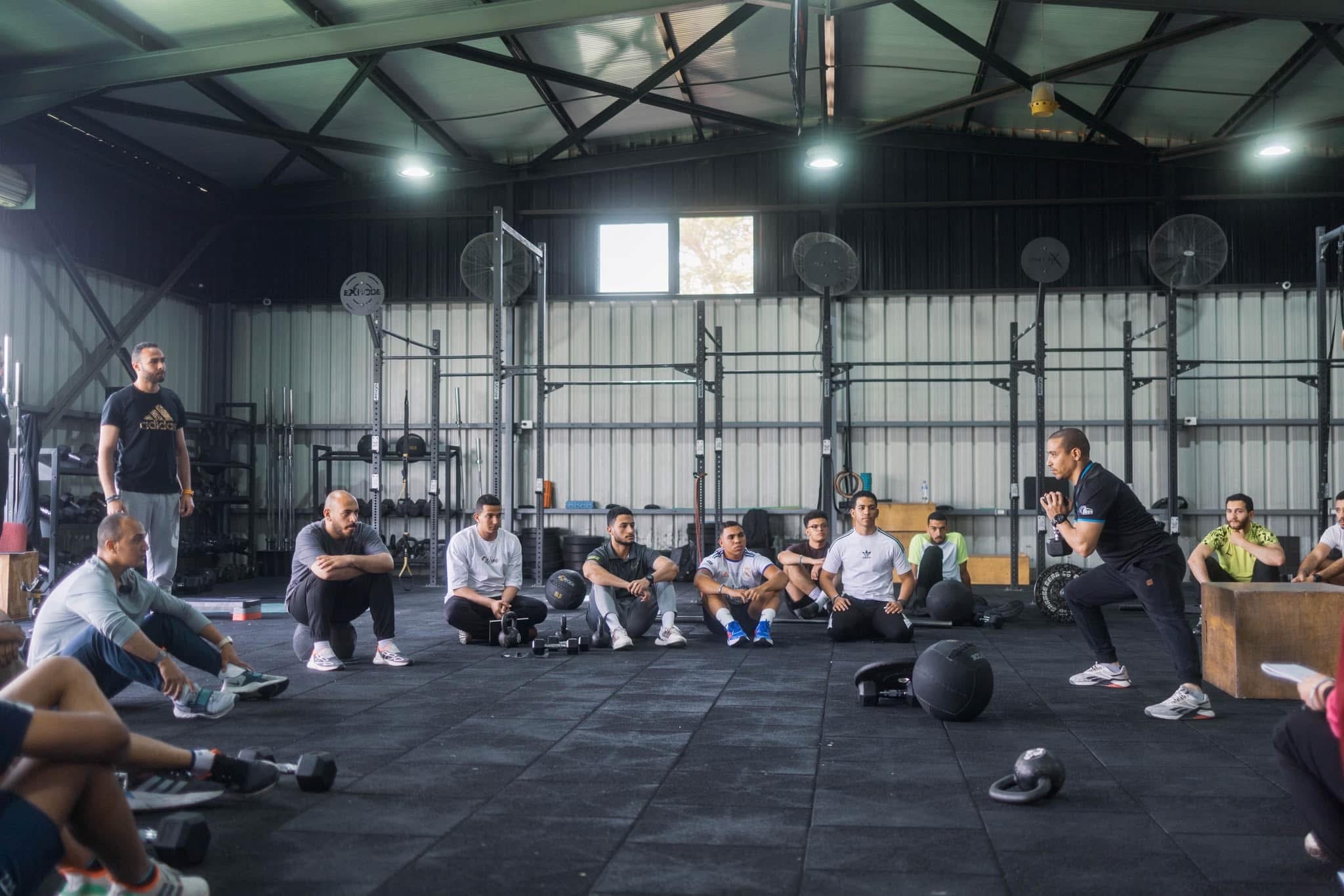 A JAHIZ coach demonstrating a loaded hinge while a squad of athletes watches from the training floor.